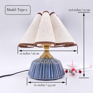 Könnte beinhalten: Eine blaue Keramik-Tischlampe mit beigem Stoffschirm und brauner Einfassung. Der Lampensockel ist 15,5 cm breit, der Schirm 26 cm breit und die Lampe 27 cm hoch. Die Modellnummer ist T050-2.