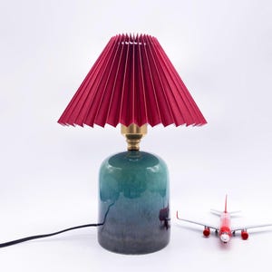May include: A decorative table lamp with a pleated burgundy lampshade and a brass neck. The base is a gradient of teal to dark blue. A small red and white toy airplane is next to the lamp.