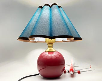 Könnte beinhalten: Eine dekorative Tischlampe mit rotem kugelförmigem Sockel und blauem Lampenschirm mit braunem Rand. Die Lampe hat einen messingfarbenen Hals. Ein Spielzeugflugzeug befindet sich neben der Lampe.