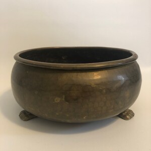 May include: A bronze-colored, round bowl with a wide rim and three claw-shaped feet. The bowl has a dark interior and a slightly textured surface. The overall design suggests a vintage or antique aesthetic, suitable for decorative purposes.
