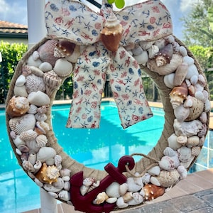 May include: A nautical-themed wreath adorned with various seashells, a burlap-wrapped base, and a decorative bow featuring anchors, starfish, and lobster patterns. A dark red anchor is attached to the bottom of the wreath. The wreath is hanging in front of a swimming pool.
