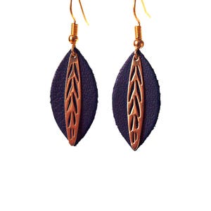Può includere: Un paio di orecchini fatti a mano con un supporto in pelle blu scuro a forma di foglia e un inserto in metallo traforato color rame. Gli orecchini hanno ganci per le orecchie color oro e una finitura strutturata, su sfondo bianco.
