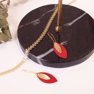 May include: Gold-colored leaf-shaped necklace and earrings. The earrings feature a red, teardrop-shaped base with a gold accent. The necklace and earrings are displayed on a black marble surface with a gold stand.