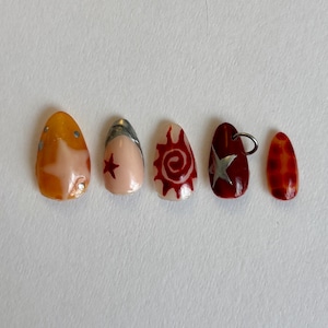 Puede incluir: Cinco uñas artificiales en forma de almendra con varios diseños. Una uña es naranja con tachuelas plateadas, otra es rosa claro con una estrella roja, y una tercera presenta un diseño de sol rojo. Una cuarta uña es rojo oscuro con una estrella plateada y un anillo de metal, y la última es roja con un patrón naranja.