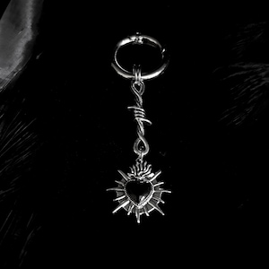 May include: A silver-toned earring with a hoop, barbed wire design, and a heart pendant. The heart is black and surrounded by a sunburst design. The earring is set against a black background.