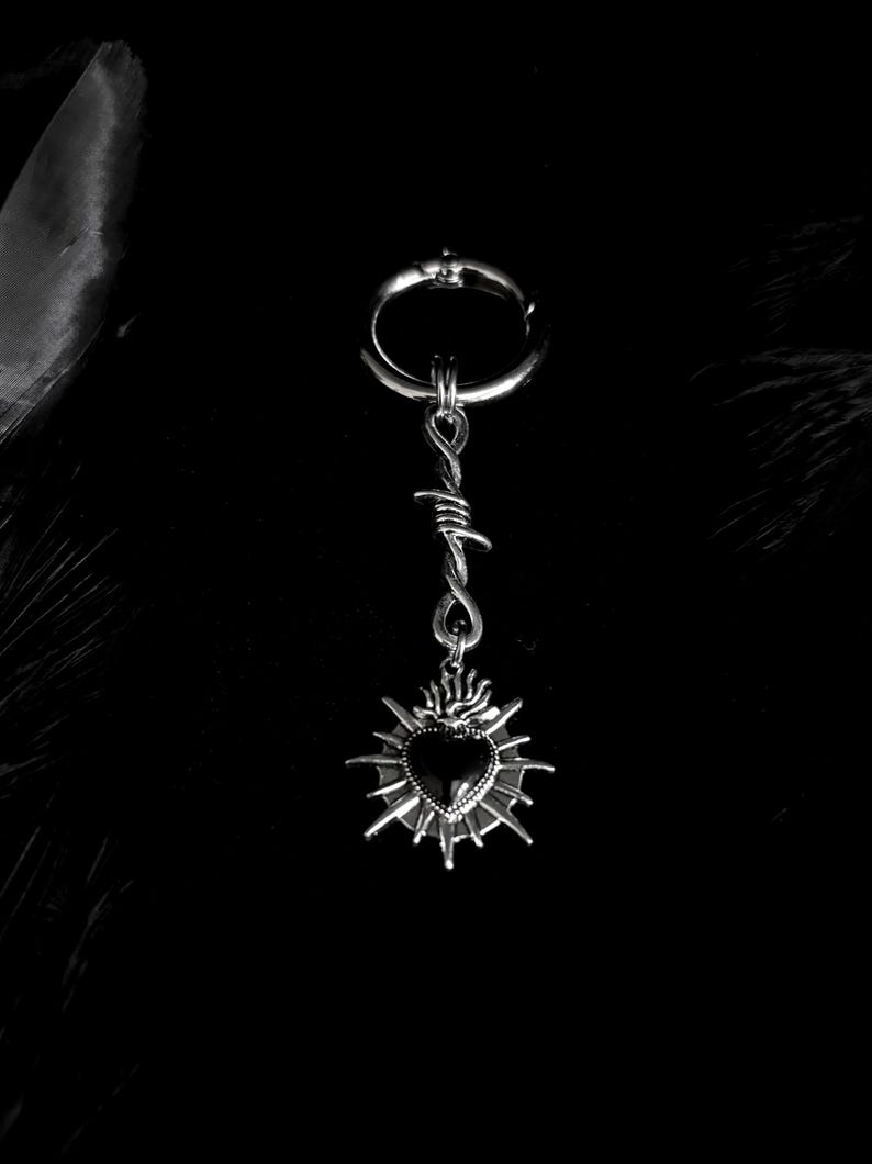 May include: A silver-toned earring with a hoop, barbed wire design, and a heart pendant. The heart is black and surrounded by a sunburst design. The earring is set against a black background.