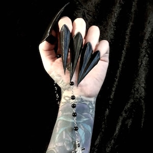 May include: Black, pointed nail covers worn on fingers. A silver rosary with black beads and a crucifix hangs from the hand. The arm has a tattoo. The background is black.