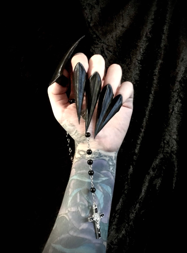 May include: Black, pointed nail covers worn on fingers. A silver rosary with black beads and a crucifix hangs from the hand. The arm has a tattoo. The background is black.