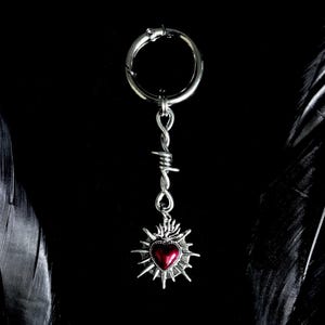 May include: A silver keychain with a barbed wire design and a pendant featuring a red heart surrounded by a sunburst design. The keychain is suspended against a black background, with a feather on either side.
