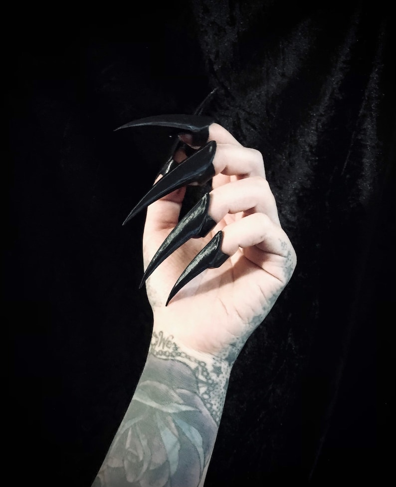 May include: A hand with long, black, pointed fingernails. The nails are sharply curved and extend beyond the fingertips. The background is a dark, solid colour, creating a dramatic contrast with the hand and nails.