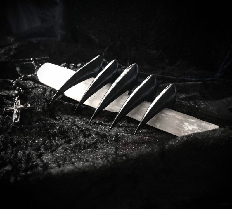 May include: A monochrome image featuring five claw-shaped objects, positioned on a white rectangular stone. A rosary with a crucifix is visible in the background, set against a dark, textured backdrop.
