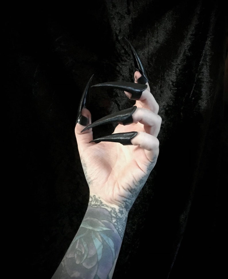 May include: A hand with long, black, pointed fingernails. The nails are glossy and extend far beyond the fingertips. The arm has a tattoo of a rose and other designs. The background is solid black.