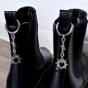 May include: A pair of black ankle boots with silver-toned charms. Each charm features a barbed wire design and a heart-shaped sunburst pendant. The boots have a pull tab with a silver ring.
