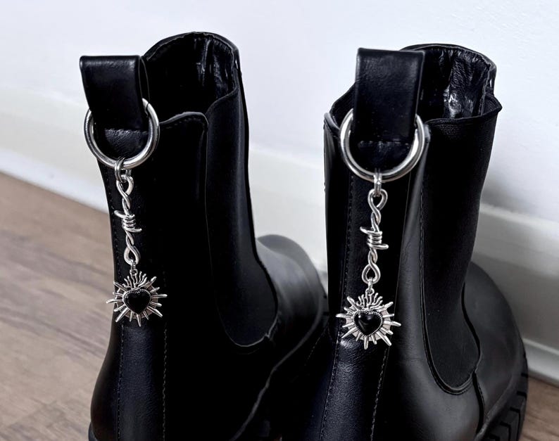 May include: A pair of black ankle boots with silver-toned charms. Each charm features a barbed wire design and a heart-shaped sunburst pendant. The boots have a pull tab with a silver ring.