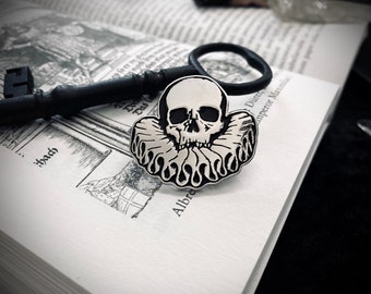 May include: A silver skull pin with a black ruff collar. The pin is on a white book with black text.