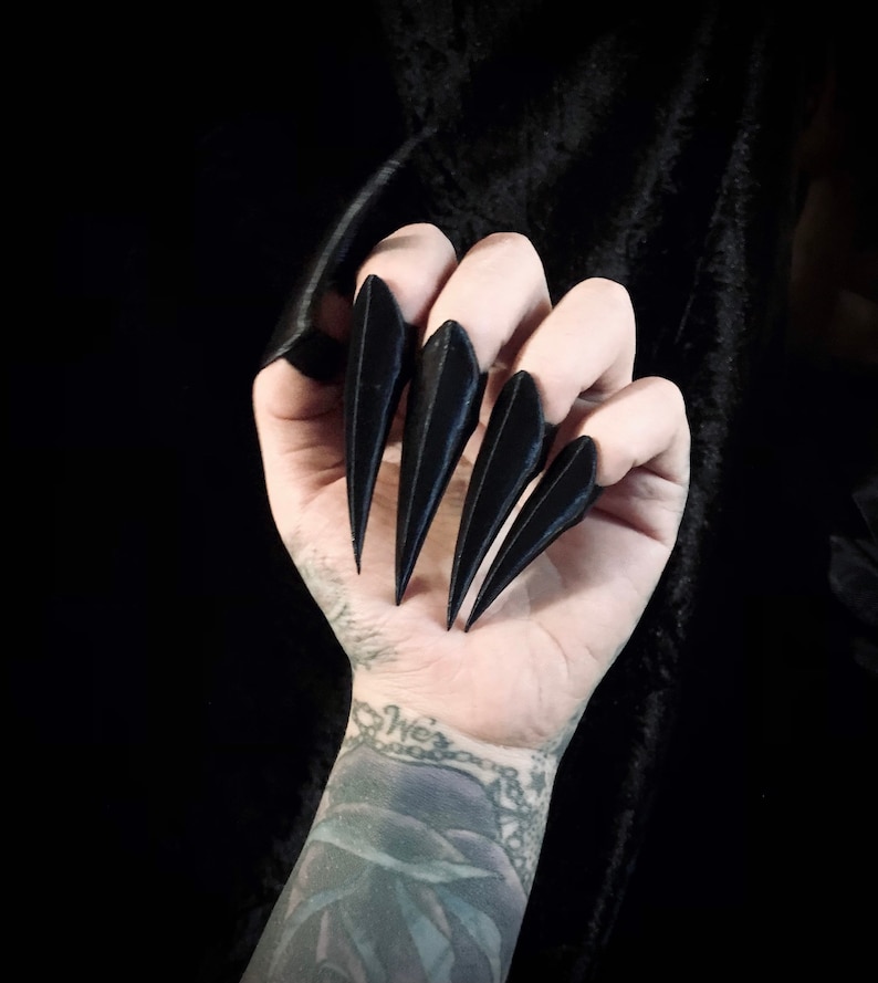 May include: A hand with black, pointed claw-shaped nail covers. The nails are long and sharply angled, creating a dramatic, gothic look. The background is a dark, solid colour, highlighting the claws.