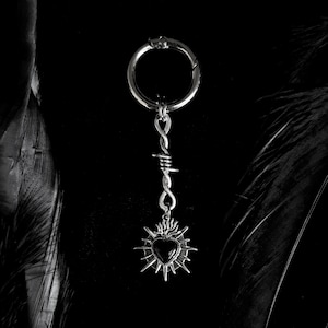 May include: A silver-colored keychain with a black heart pendant. The pendant is surrounded by a sunburst design and attached to a barbed wire-style chain. The keychain has a circular clasp.