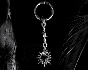 May include: A silver-colored keychain with a black heart pendant. The pendant is surrounded by a sunburst design and attached to a barbed wire-style chain. The keychain has a circular clasp.