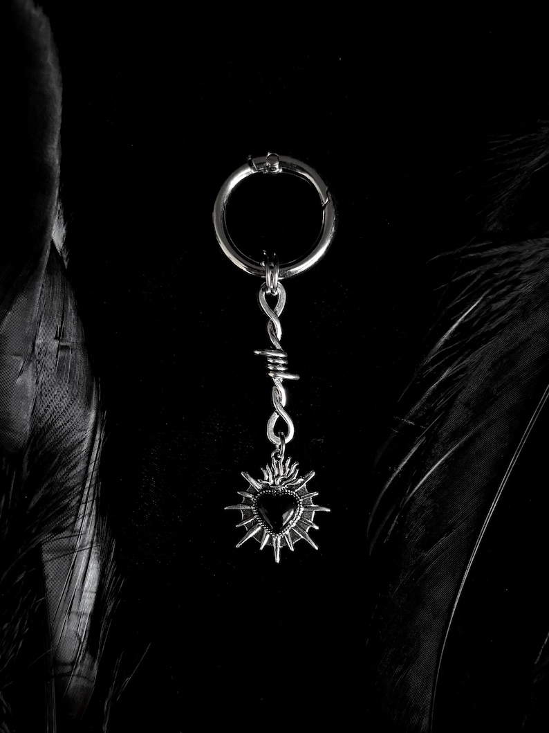 May include: A silver-colored keychain with a black heart pendant. The pendant is surrounded by a sunburst design and attached to a barbed wire-style chain. The keychain has a circular clasp.