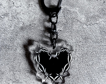 May include: Black heart-shaped keychain with a barbed wire design. The keychain has a black clasp and chain. The heart is filled with black and outlined with white barbed wire. The background is a textured gray surface.