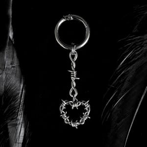 May include: A silver-colored keychain with a heart-shaped barbed wire pendant. The keychain has a circular clasp at the top and is suspended from a series of small loops. The background is black.
