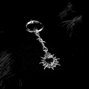 May include: A silver-toned earring with a barbed wire design and a pendant featuring a black heart surrounded by flames. The earring has a hoop closure and a dark background.