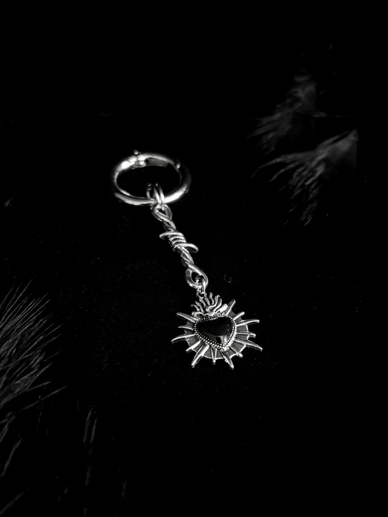 May include: A silver-toned earring with a barbed wire design and a pendant featuring a black heart surrounded by flames. The earring has a hoop closure and a dark background.