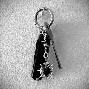 May include: A black and silver keychain with a Swiss Army knife, keys, and a heart-shaped charm. The charm has a dark center and a sunburst design. The keychain is attached to a silver ring.