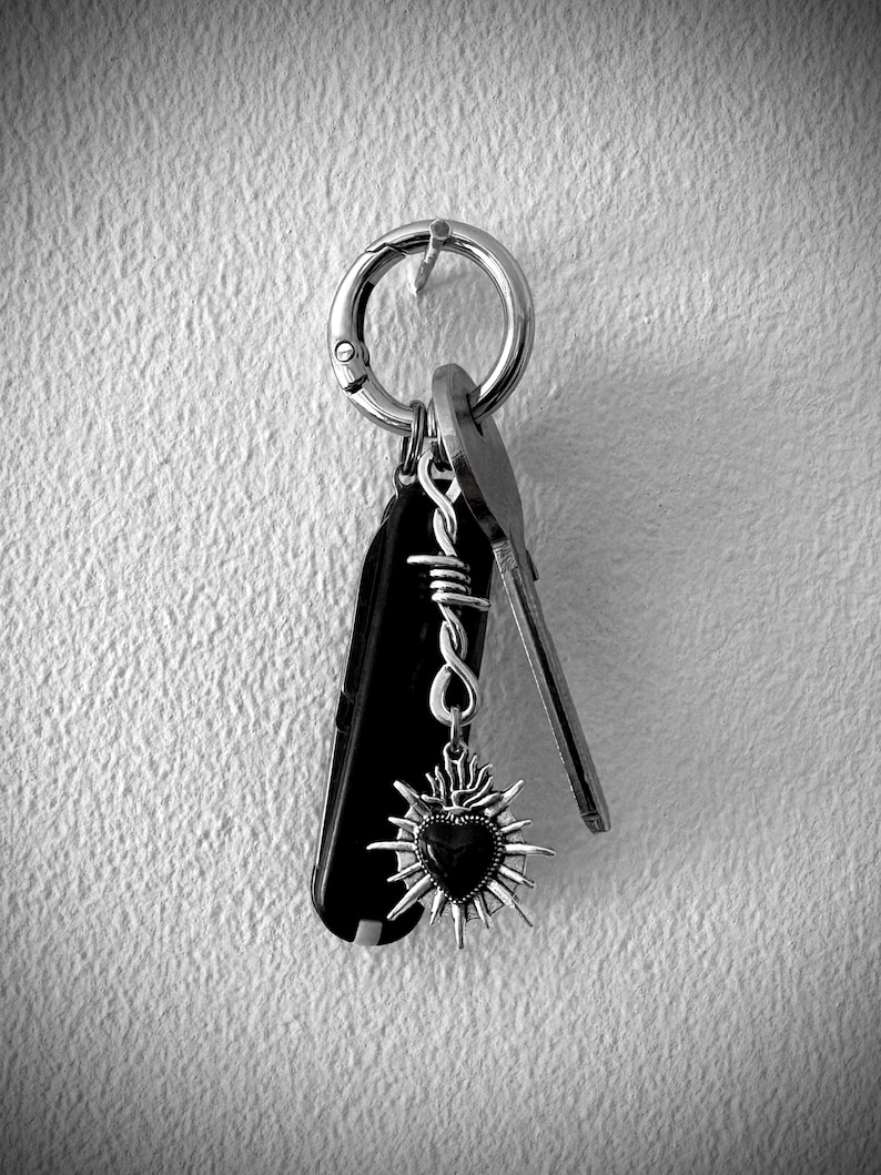 May include: A black and silver keychain with a Swiss Army knife, keys, and a heart-shaped charm. The charm has a dark center and a sunburst design. The keychain is attached to a silver ring.