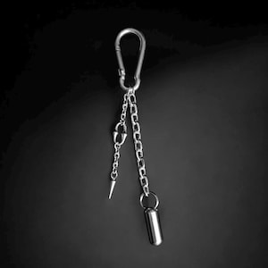 Może przedstawiać: Srebrny brelok do kluczy z karabińczykiem, dwoma łańcuszkami, kolcem i cylindrycznym wisiorkiem. Łańcuszki zwisają z karabińczyka, z kolcem i wisiorkiem zawieszonymi na końcach. Tło jest jednolicie czarne.