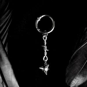 Può includere: Un orecchino argentato con un cerchio circolare e un ciondolo pendente. Il ciondolo presenta un design a filo ritorto e un pendente a forma di stella appuntita. L'orecchino è posto su uno sfondo scuro, con elementi a forma di piuma su entrambi i lati.