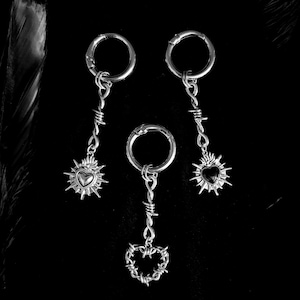 May include: Three silver-toned earrings with a gothic aesthetic. Each earring features a circular clasp, a barbed wire design, and a heart pendant. The heart pendants have a sunburst design. The background is black.