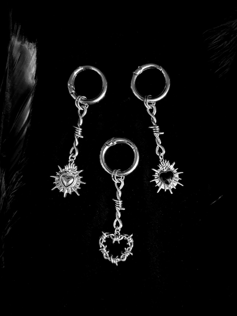 May include: Three silver-toned earrings with a gothic aesthetic. Each earring features a circular clasp, a barbed wire design, and a heart pendant. The heart pendants have a sunburst design. The background is black.