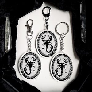 May include: Three black and white scorpion keychains with silver chains. Each keychain features a scorpion design with a black and white mandala pattern.