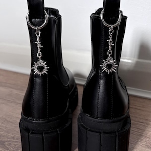 May include: Pair of black platform boots with silver-tone charms. The boots have a chunky sole and a pull tab at the back. Each boot features a dangling charm with a heart and barbed wire design.