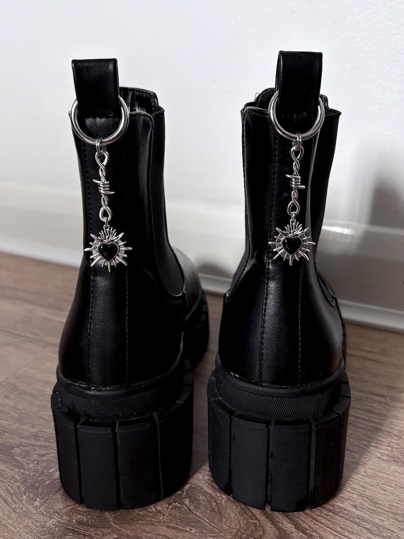 May include: Pair of black platform boots with silver-tone charms. The boots have a chunky sole and a pull tab at the back. Each boot features a dangling charm with a heart and barbed wire design.