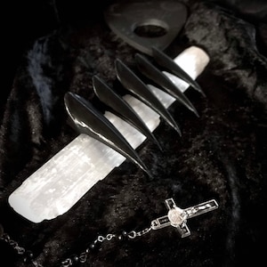 May include: Black claw-shaped nail covers are displayed on a white crystal, with a rosary and a planchette in the background. The items are set against a dark, textured backdrop, creating a gothic aesthetic.