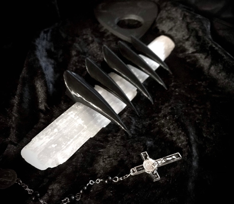May include: Black claw-shaped nail covers are displayed on a white crystal, with a rosary and a planchette in the background. The items are set against a dark, textured backdrop, creating a gothic aesthetic.