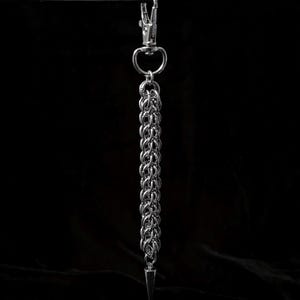 Peut inclure: Un porte-clés en cotte de mailles argentée avec un mousqueton et un pendentif en forme de pointe conique. La cotte de mailles est faite de petits anneaux imbriqués. Le fond est noir.