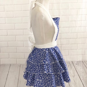 May include: A blue and white apron with a snowflake pattern. The apron has a white halter strap and a ruffled skirt.