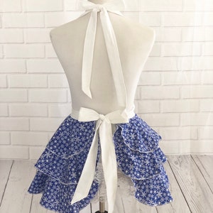 May include: A blue and white snowflake print apron with a white ruffled skirt and white straps. The apron is tied at the back with a bow.