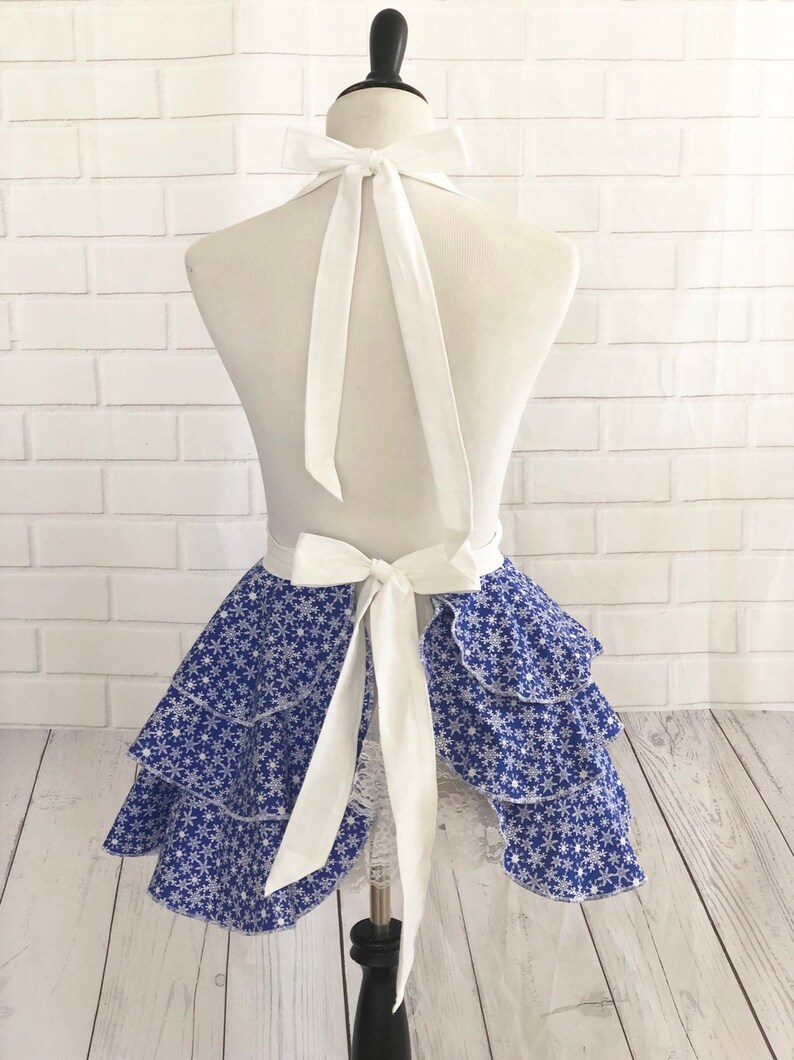 May include: A blue and white snowflake print apron with a white ruffled skirt and white straps. The apron is tied at the back with a bow.