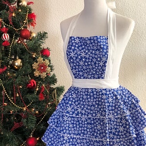 May include: A blue and white snowflake patterned apron with a white ruffle skirt and white halter straps. The apron is on a white mannequin.