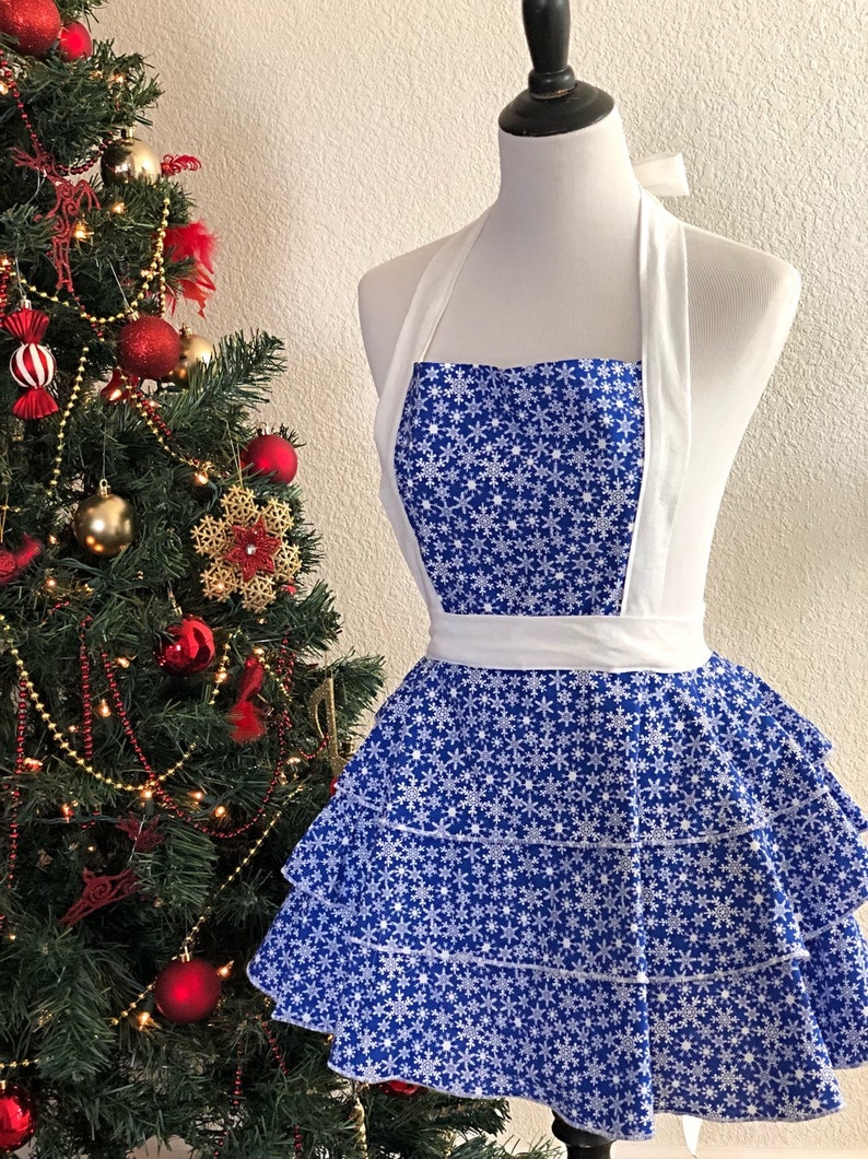 May include: A blue and white snowflake patterned apron with a white ruffle skirt and white halter straps. The apron is on a white mannequin.
