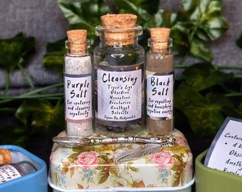 May include: Three small glass bottles with cork stoppers containing purple, brown, and clear crystals. The bottles are labeled "Purple Salt", "Cleansing", and "Black Salt". The bottles are sitting on a small tin box with a floral pattern. The box also contains a small silver feather.