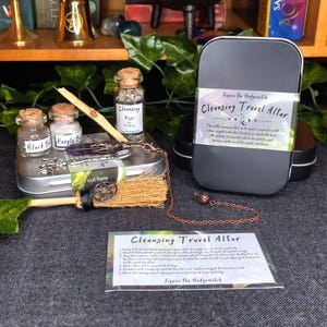 May include: A cleansing travel altar kit featuring a black tin, small glass bottles, a miniature broom, and a copper chain. The kit includes bottles labeled "Black Salt", "Purple Salt", and "Cleansing Sage". The text "Cleansing Travel Altar" is visible.