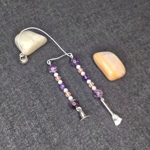Puede incluir: Un par de pendientes colgantes con un dije de sombrero de bruja y escoba de plata. Los pendientes están hechos con cuentas de amatista y rosa.
