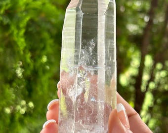 May include: A clear quartz crystal point with a faceted top. The crystal is held against a blurred green background. The quartz is transparent with visible internal structures and a rough base.
