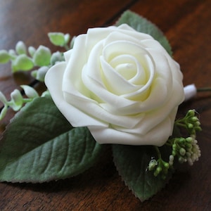 May include: A single white rose with green leaves and small white buds. The rose is a silk flower and is part of a boutonniere.