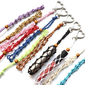 May include: A collection of colorful macrame keychains with wooden beads and heart-shaped clasps. The keychains are in various colors, including purple, brown, pink, red, blue, green, black, white, and turquoise. Some keychains hold cylindrical objects.